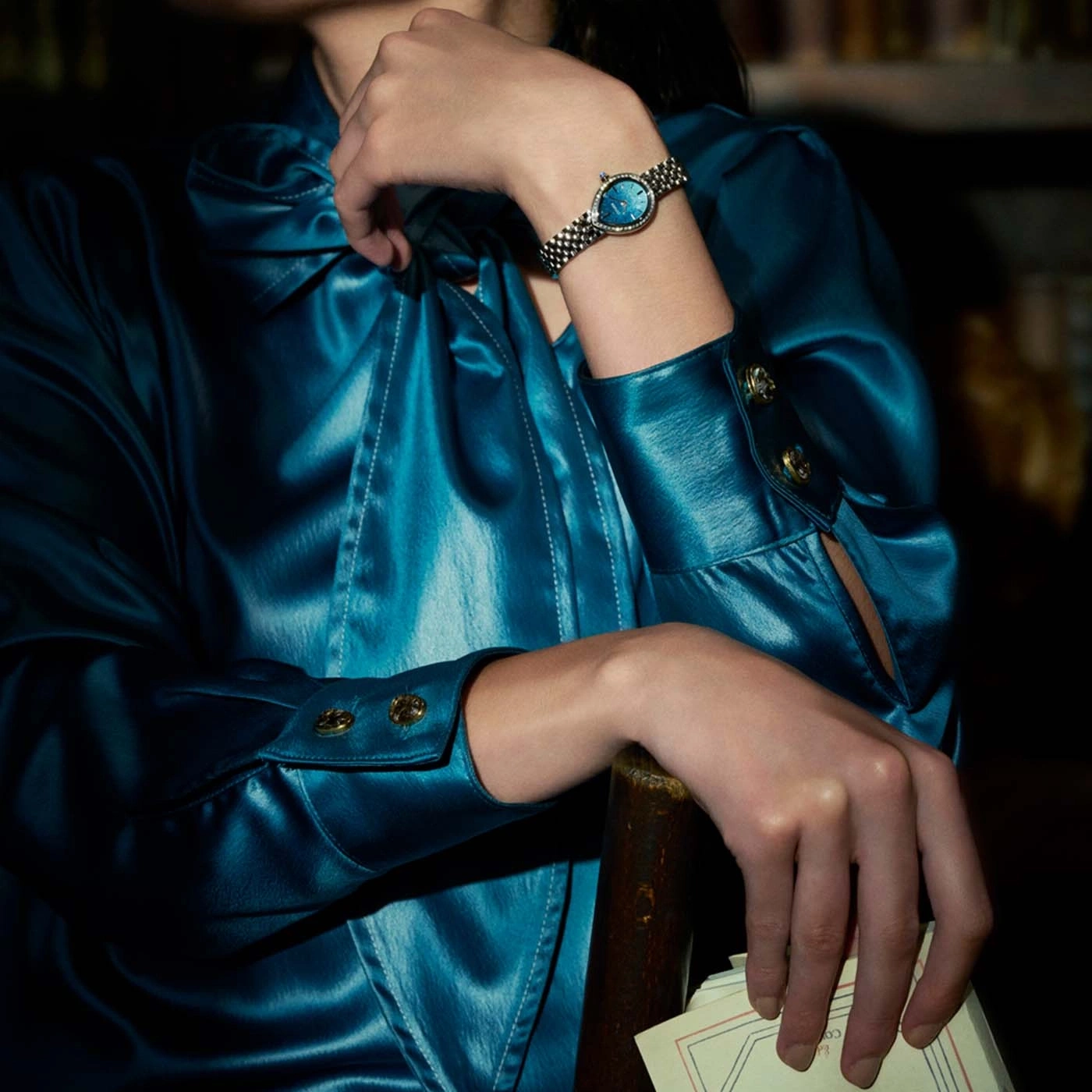 Frau in einem blauen Satin-Kleid mit einer Brille und einer Uhr am Handgelenk.