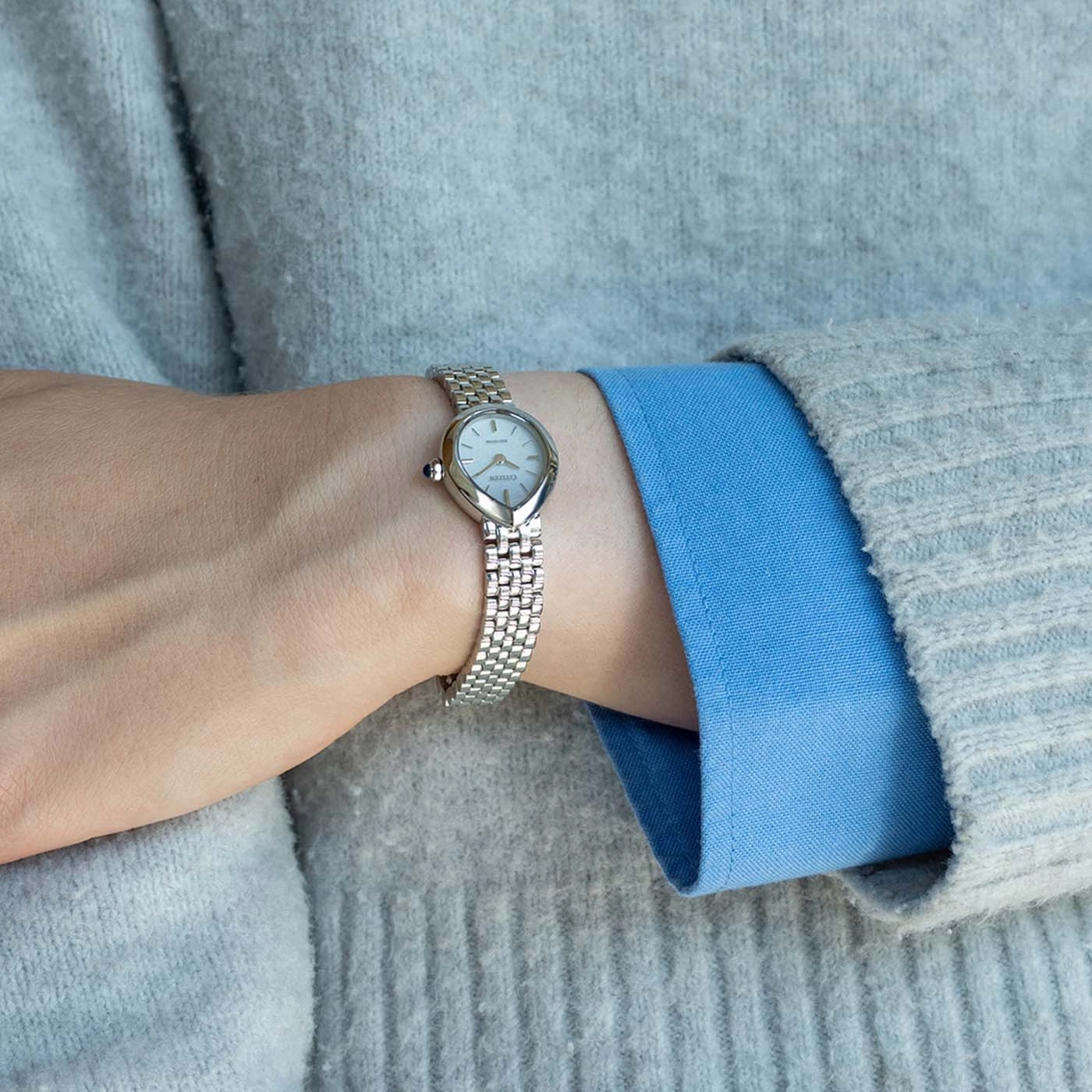 Eine Hand mit einer silbernen Armbanduhr über einem grauen Pullover und einem blauen Hemd.