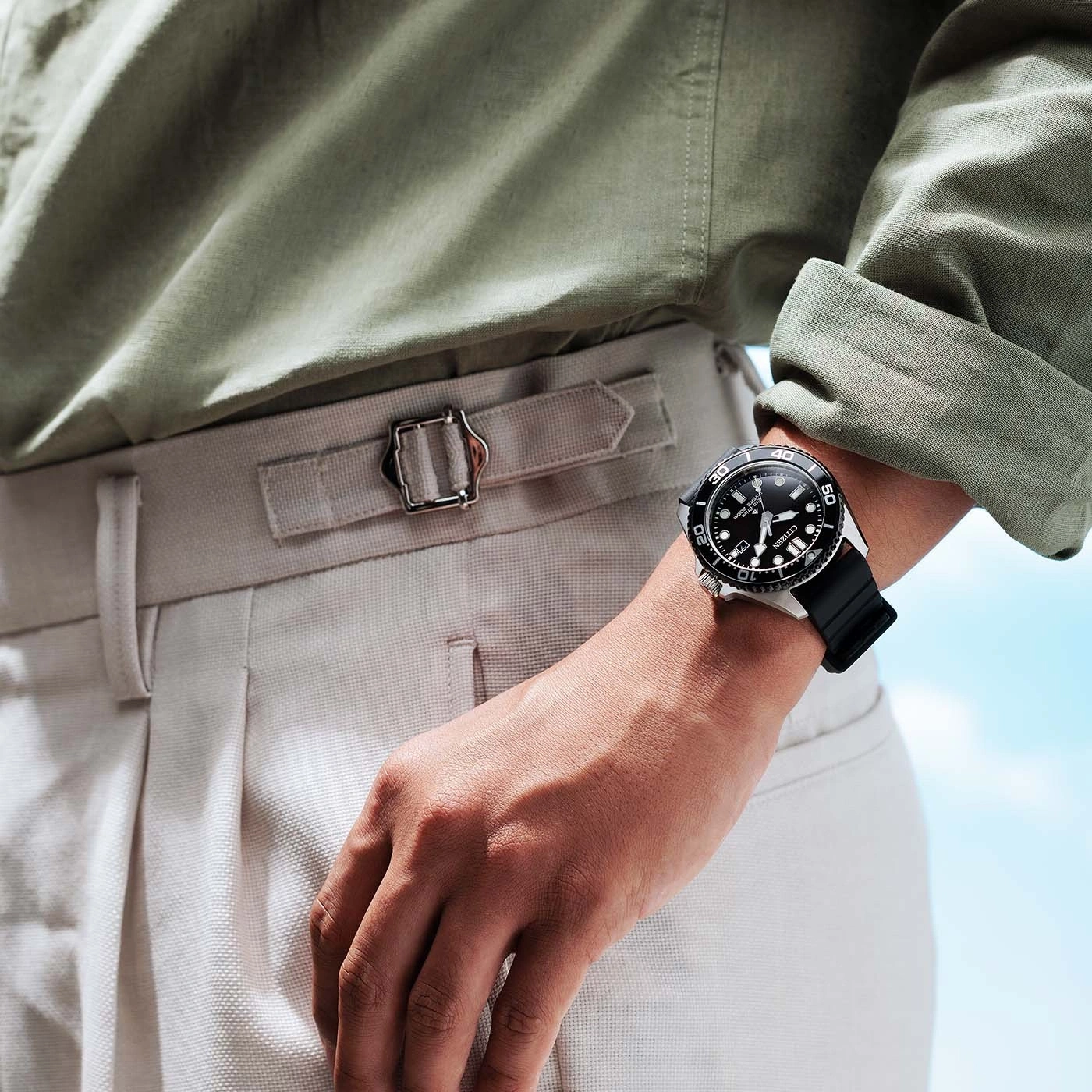 Un homme porte une chemise verte et un pantalon beige avec une montre Citizen au poignet.