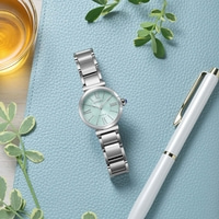 Une montre Citizen Eco-Drive vert menthe avec un bracelet argenté repose sur une surface texturée bleue à côté d'un stylo blanc et d'un verre de liquide ambré.