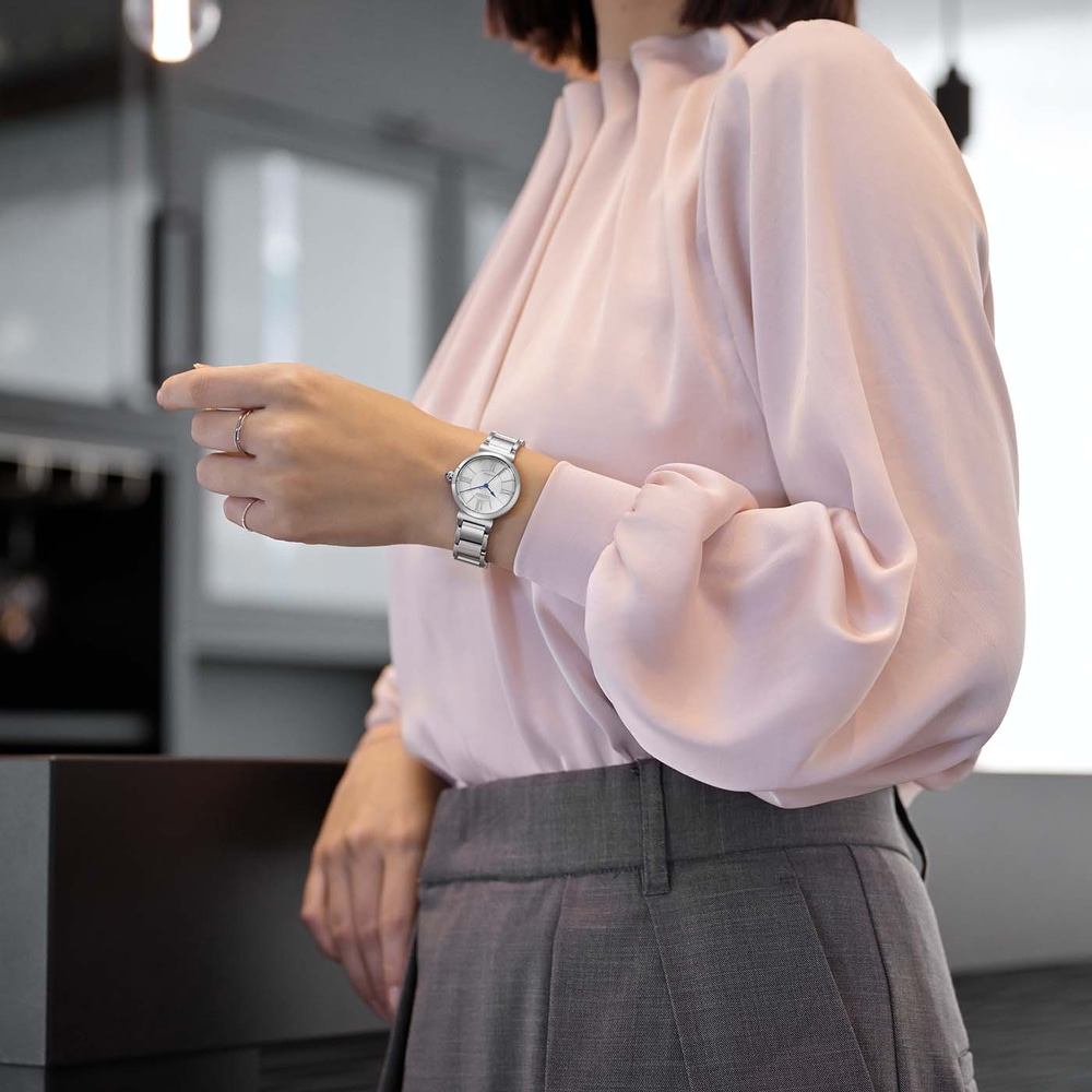 Femme portant un chemisier rose et un pantalon gris, regardant sa montre.