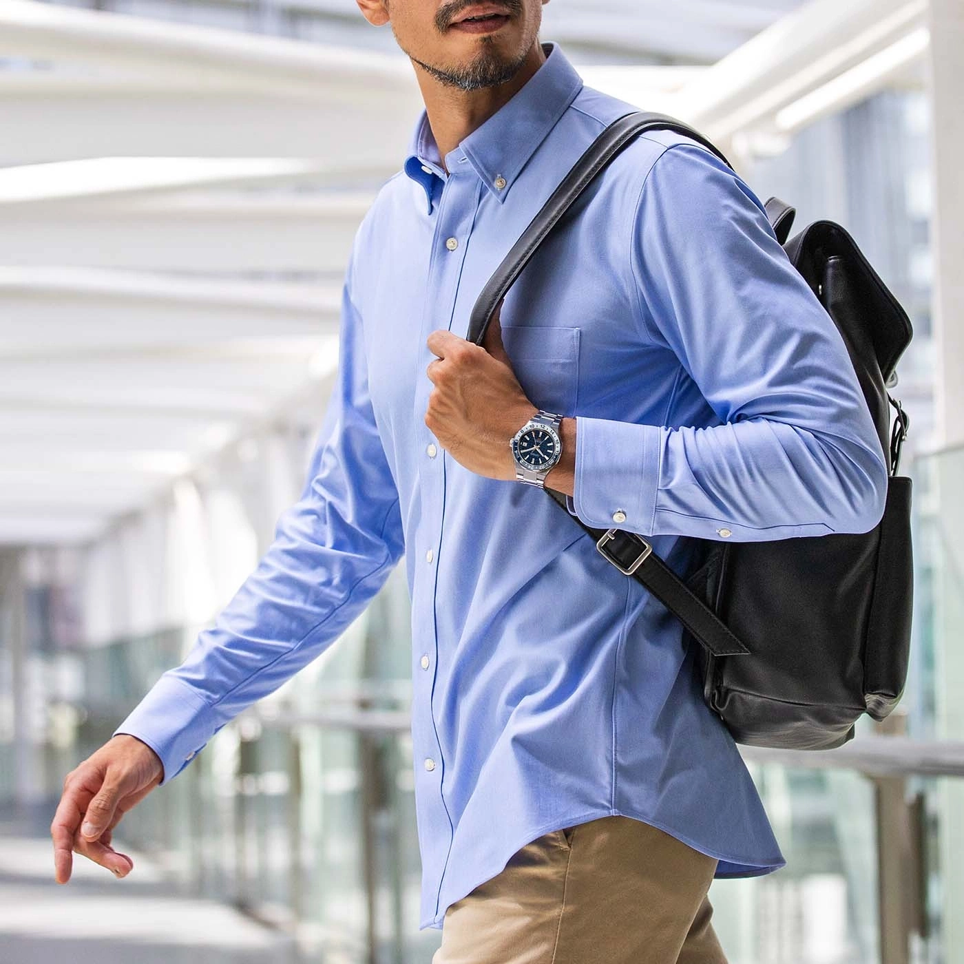Un homme en chemise bleue porte un sac à dos noir et une montre.