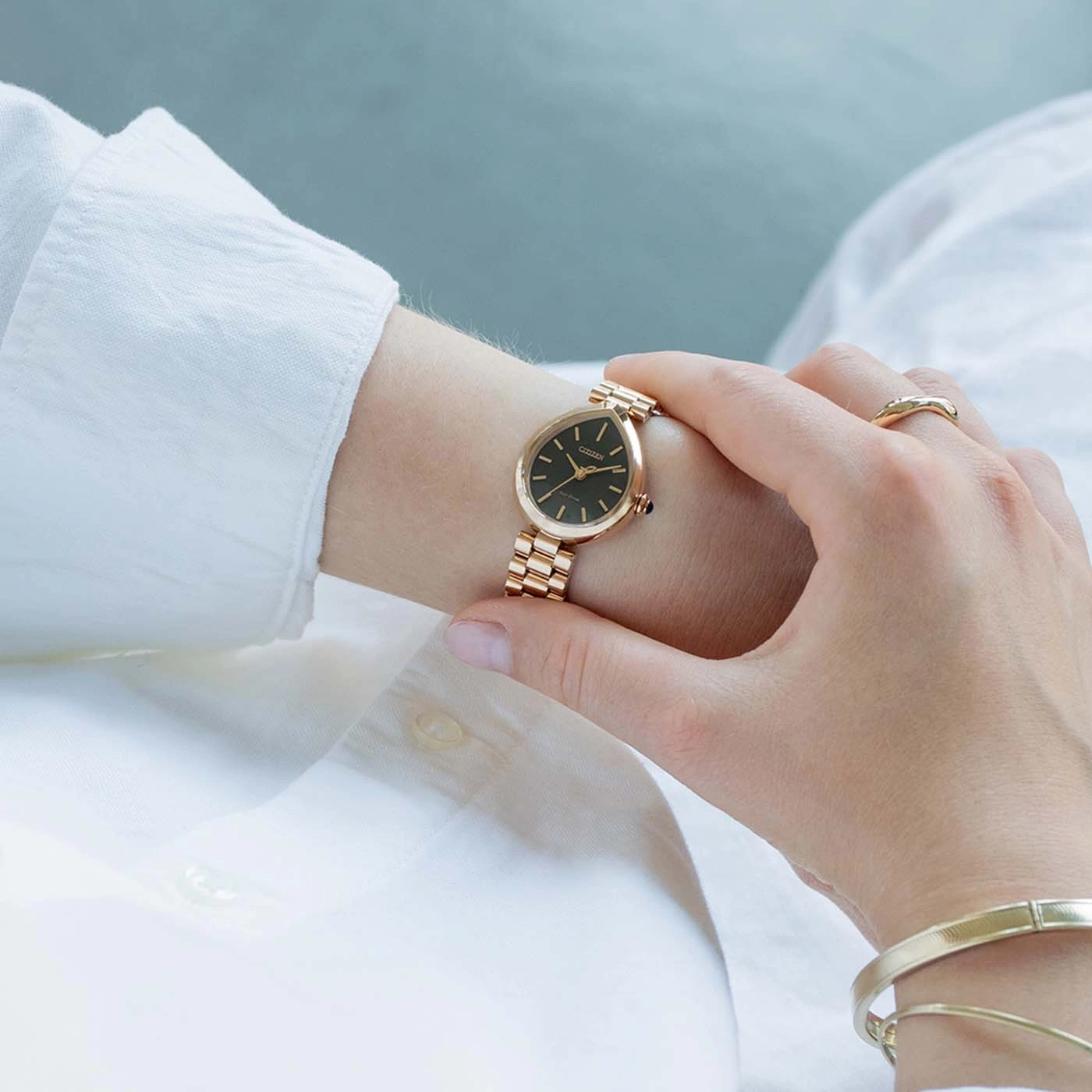 Eine Frau trägt eine goldene Citizen-Uhr am rechten Handgelenk mit einem Ring und Armband.