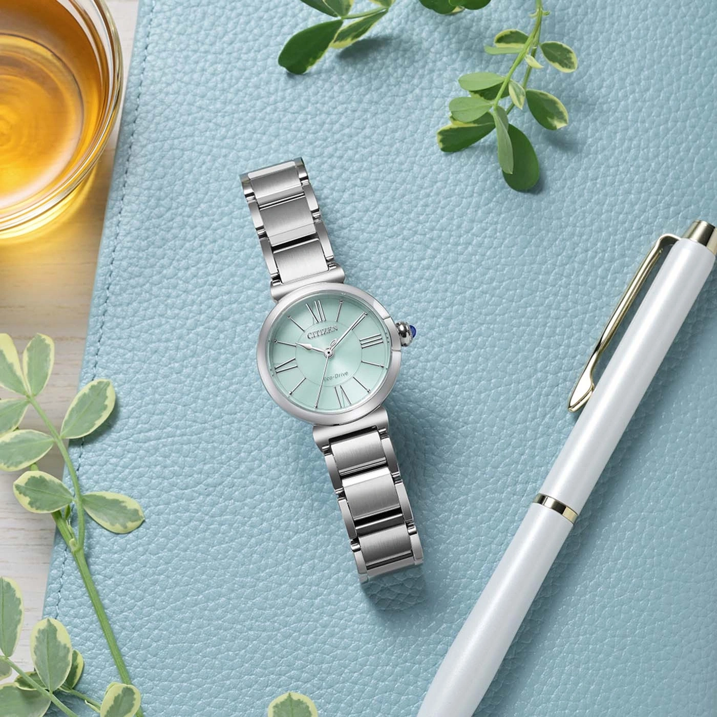 Eine mintgrüne Citizen Eco-Drive-Uhr liegt neben einem weißen Stift auf einem blaugrünen Hintergrund.