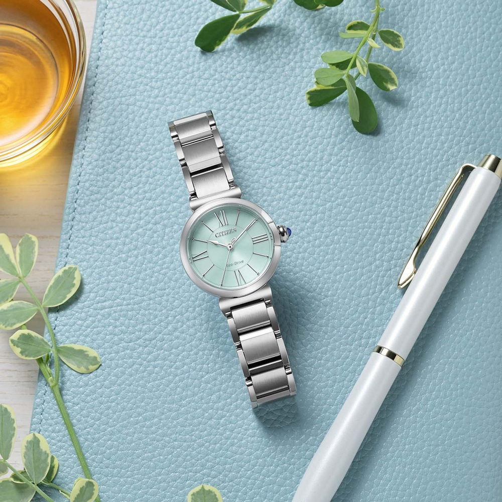 Une montre Citizen Eco-Drive vert menthe avec un bracelet argenté repose sur une surface texturée bleue à côté d'un stylo blanc et d'un verre de liquide ambré.