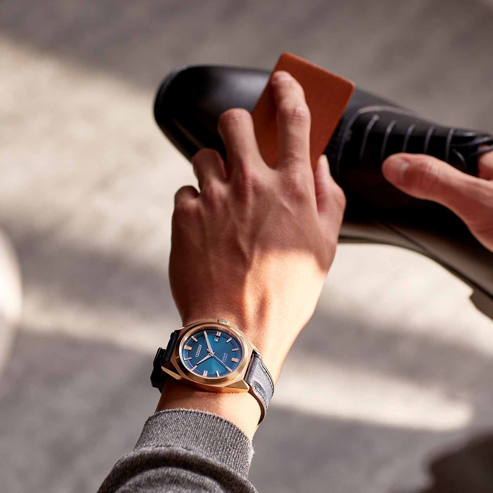 Homme polissant une chaussure noire, portant une montre dorée au cadran bleu.