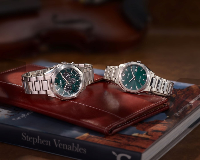 Zwei Citizen-Uhren mit grünen Zifferblättern auf einem Buch und einer Ledergeldbörse.