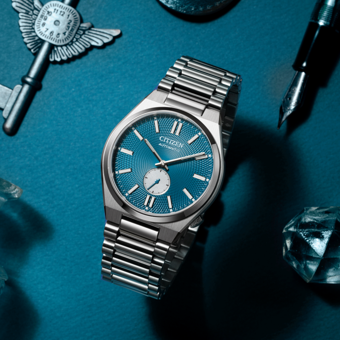 Citizen-Uhr mit blauem Zifferblatt, Schlüssel, Stift und Kristallen auf blauem Untergrund.