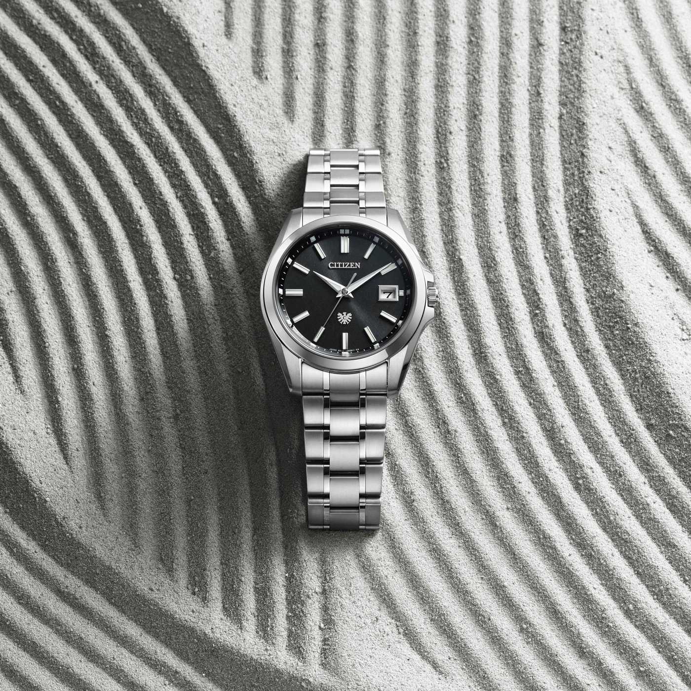 Eine silberne Citizen-Uhr liegt auf Sand mit parallelen Streifen.