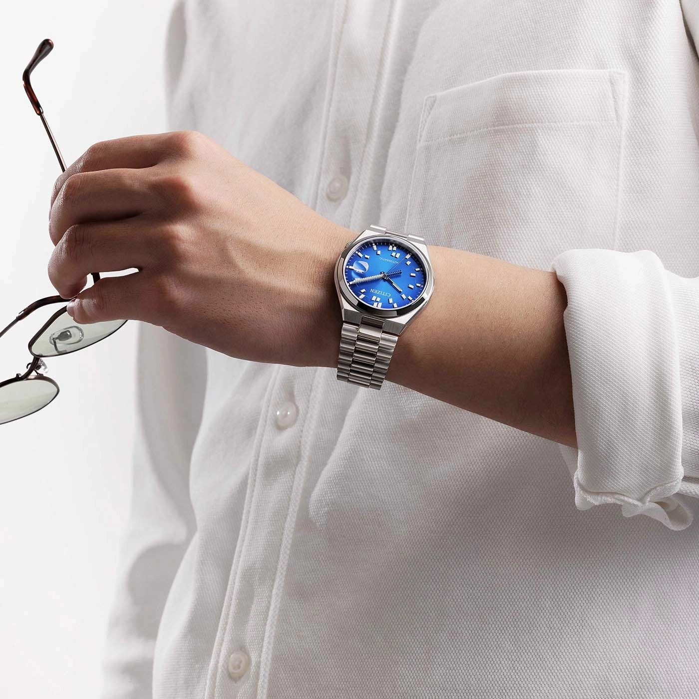 Mann hält Brille, trägt blaue Citizen-Uhr am weißen Poloshirt.