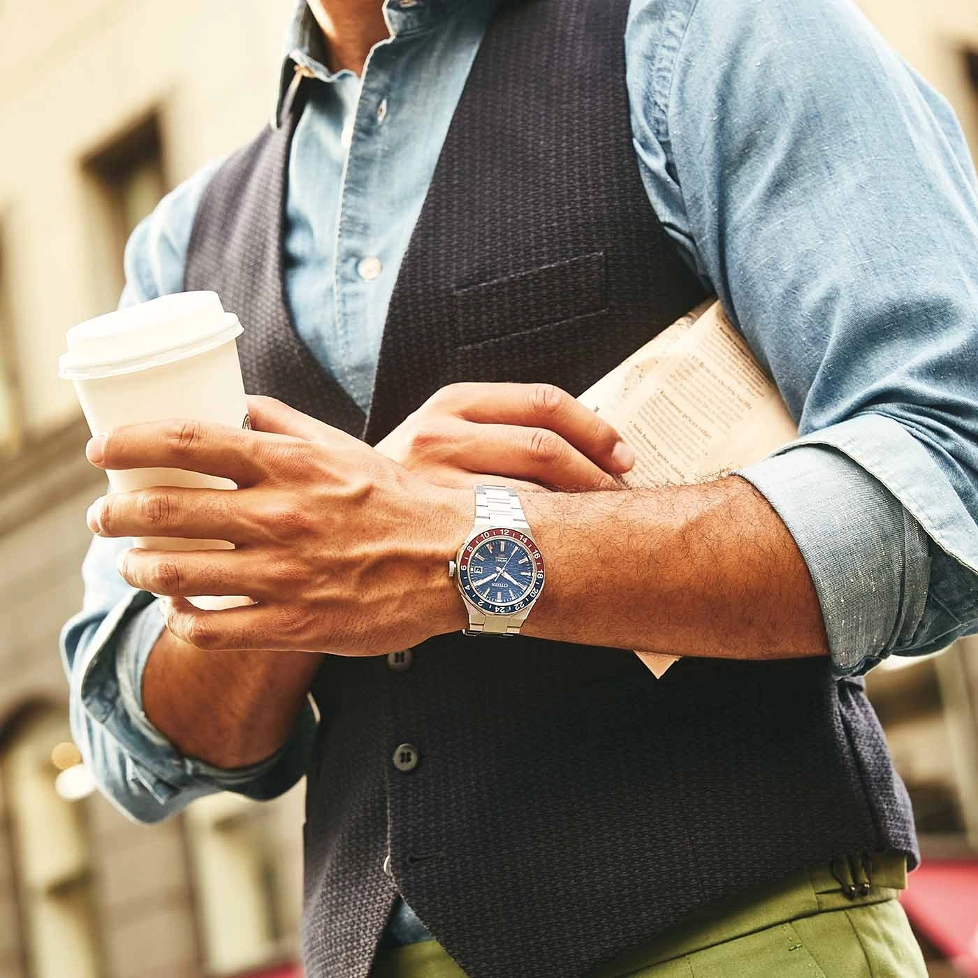 Man holding coffee and newspaper, wearing a watch and vest.