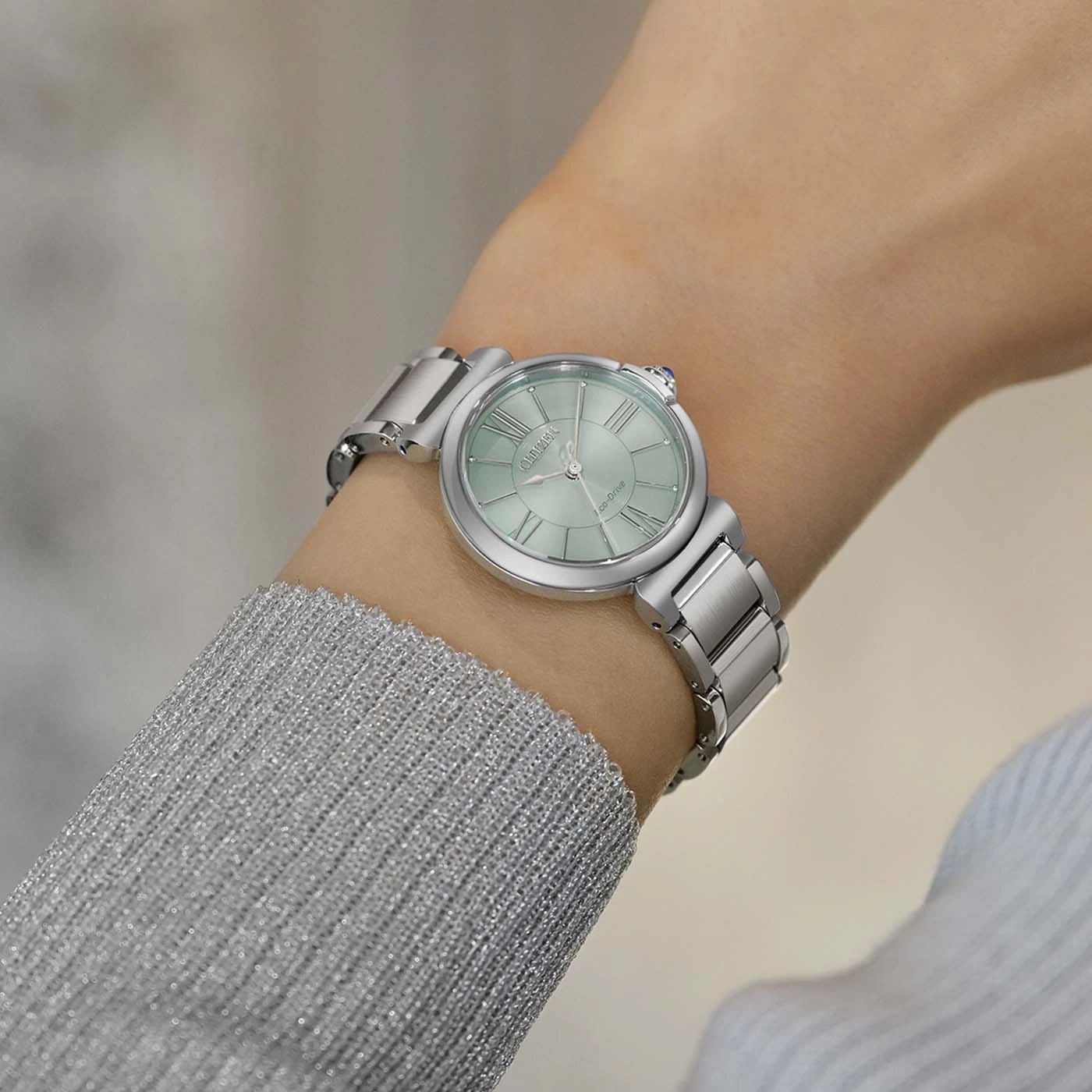 Eine Frau trägt eine silberne Uhr am linken Handgelenk, mit einem blassgrünen Zifferblatt und römischen Ziffern.