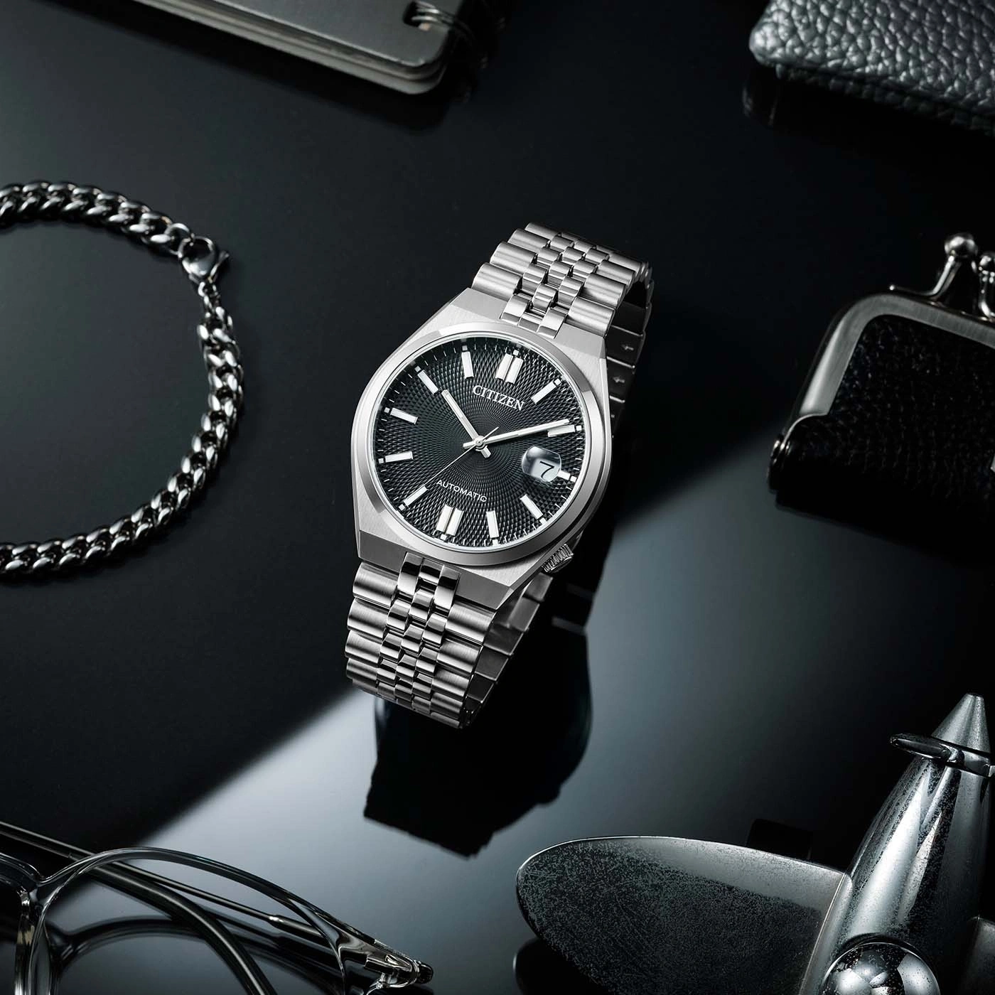 Montre Citizen avec bracelet en argent sur fond noir, entourée d'accessoires.