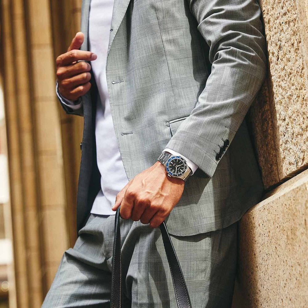 Homme en costume avec montre et sac appuyé contre un mur