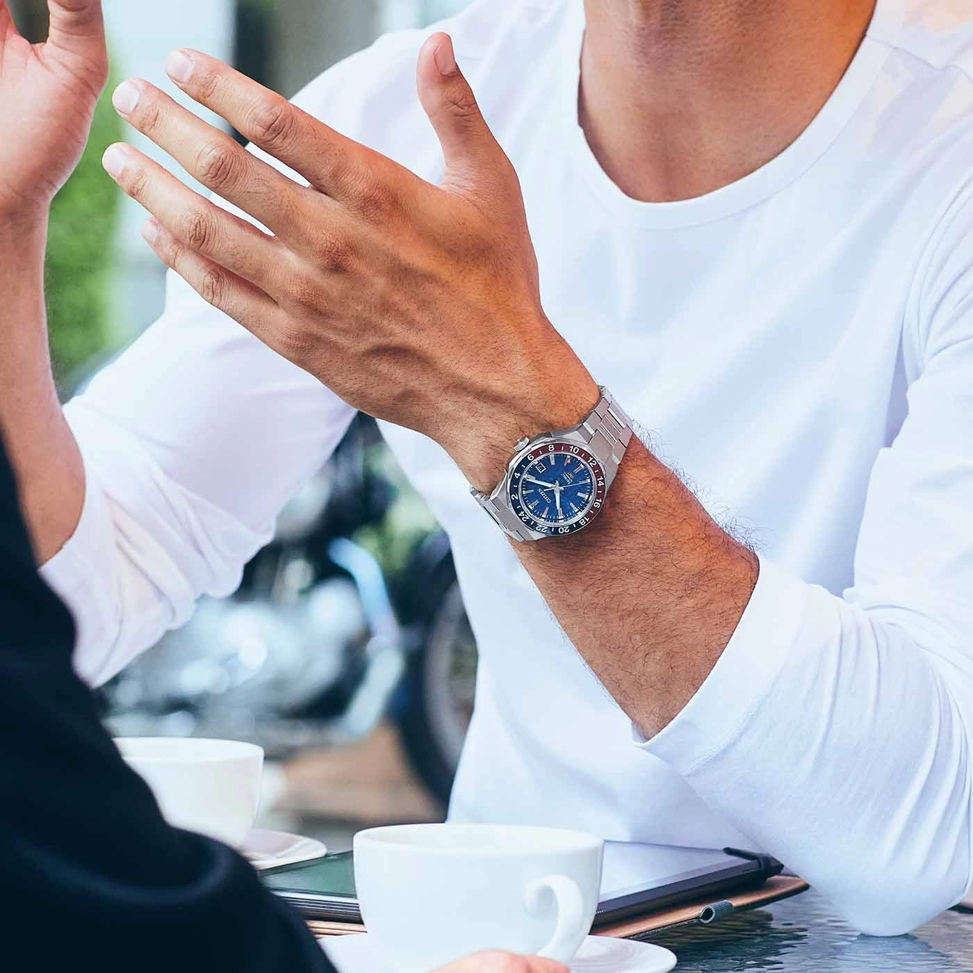 Man wearing a watch, gesturing with hands, sitting at a table with coffee cups.